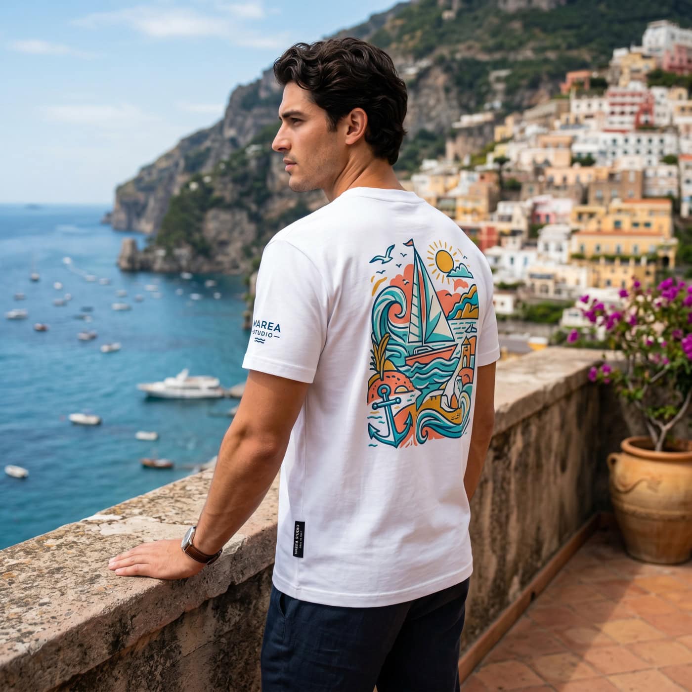 Un hombre con una camiseta blanca con un colorido diseño náutico está de pie en una terraza con vistas a un pueblo costero y al mar, donde se ven barcos.
