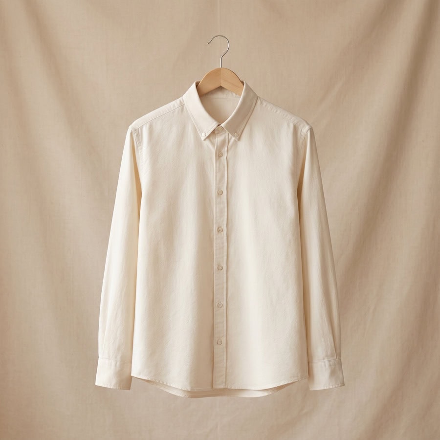 Una camisa de manga larga, de color blanco roto y con botones, colgada de una percha de madera y expuesta sobre un fondo de tela beige a juego: perfecta para quienes buscan prendas personalizadas con un estilo atemporal.