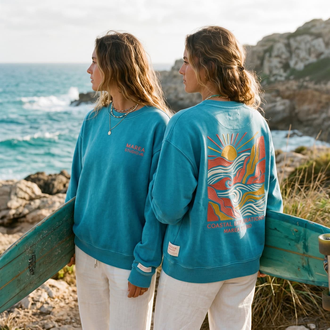 Dos personas que llevan sudaderas azules y pantalones blancos están de pie junto al mar, con una tabla de surf en las manos. Una de las sudaderas tiene una colorida ilustración costera en la espalda.